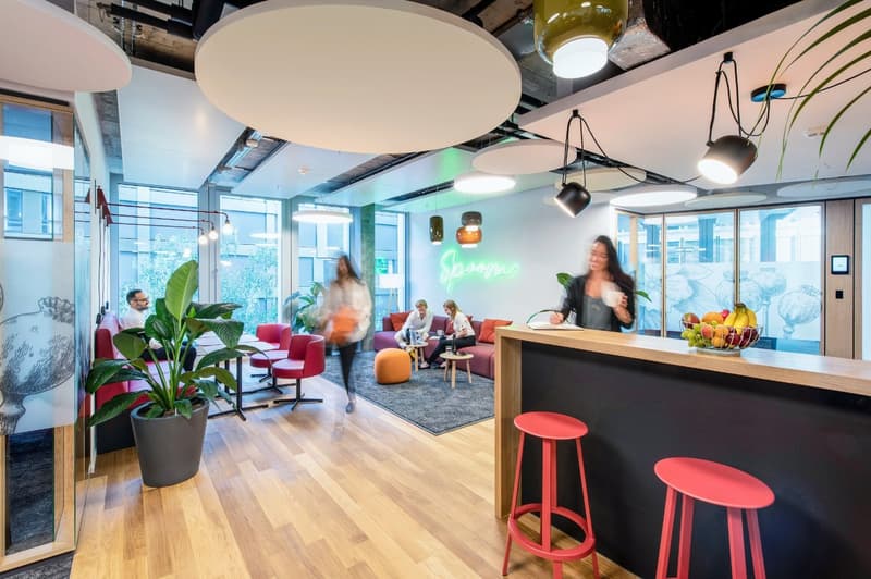Open concept office space with hardwood floors, large windows, modern lighting fixtures, potted plants, red and orange furniture, and a kitchen area with a dishwasher