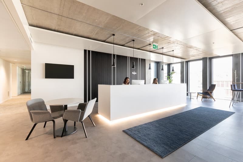 Reception area with reception desk, large monitor, chairs, desk, two people working, bright space, large windows