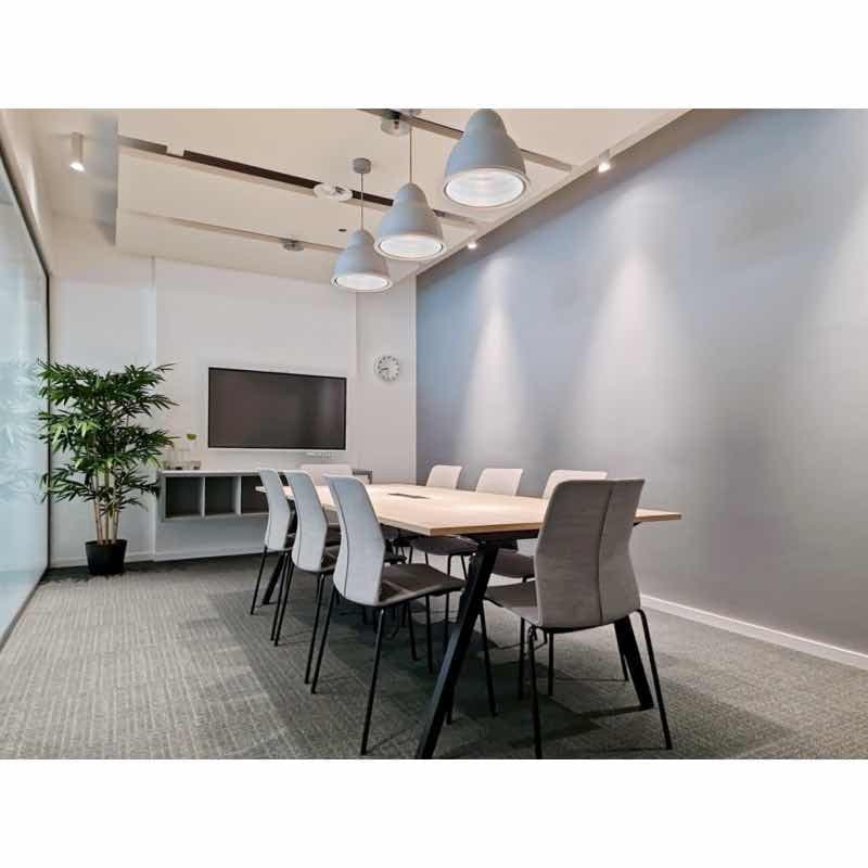 Modern meeting room with a long wooden table, gray chairs, a TV mounted on the wall, and a potted plant in the corner.