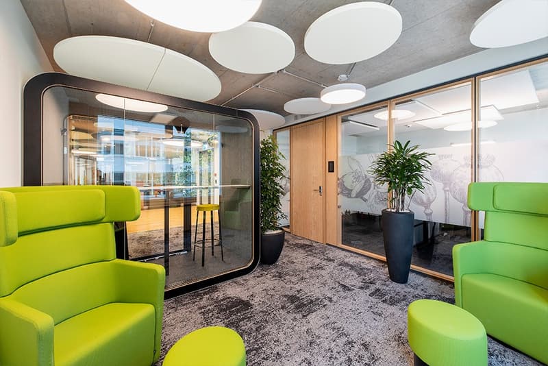 Modern office space with glass-enclosed meeting room, green upholstered chairs and ottomans, large potted plant, and circular lighting fixtures on the ceiling.