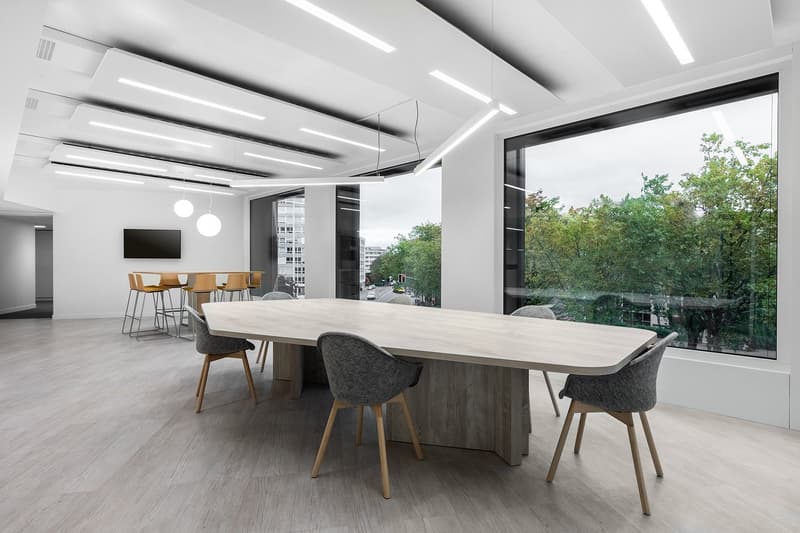 Large open office space with a long wooden conference table, gray upholstered chairs, and a large window overlooking a wooded area. The ceiling has recessed lighting fixtures and the floor is a light-colored wood laminate. There is a TV mounted on the wal
