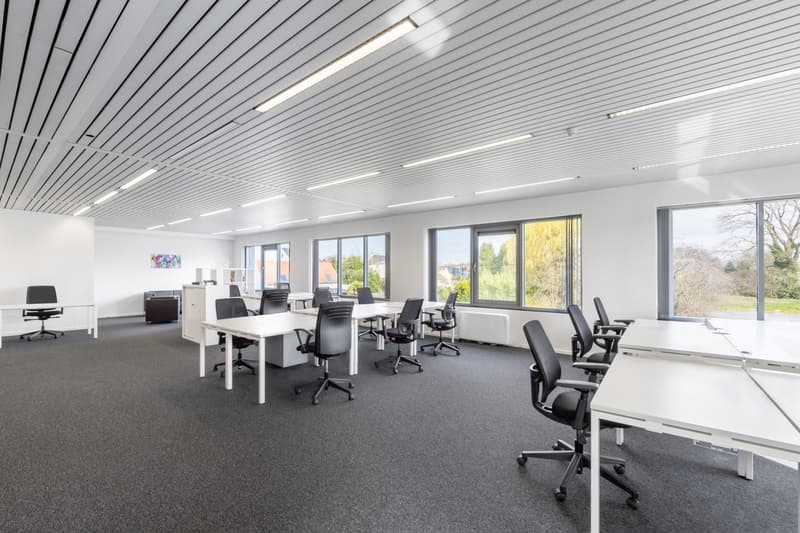 Large open office space with multiple workstations, desks, and chairs. The room has large windows providing natural light and a view of the outdoors. The ceiling is lined with fluorescent lighting fixtures. The floor is covered in dark gray carpeting.