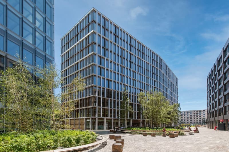 A modern, multi-story office building with a glass facade and a landscaped plaza in front. The building has a rectangular shape with a stepped design, and there are balconies or terraces visible on some of the floors.