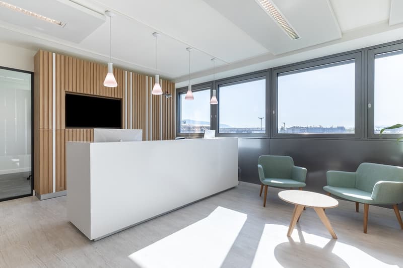reception desk, chairs, round table, large windows, monitor on wall, wood and white color scheme