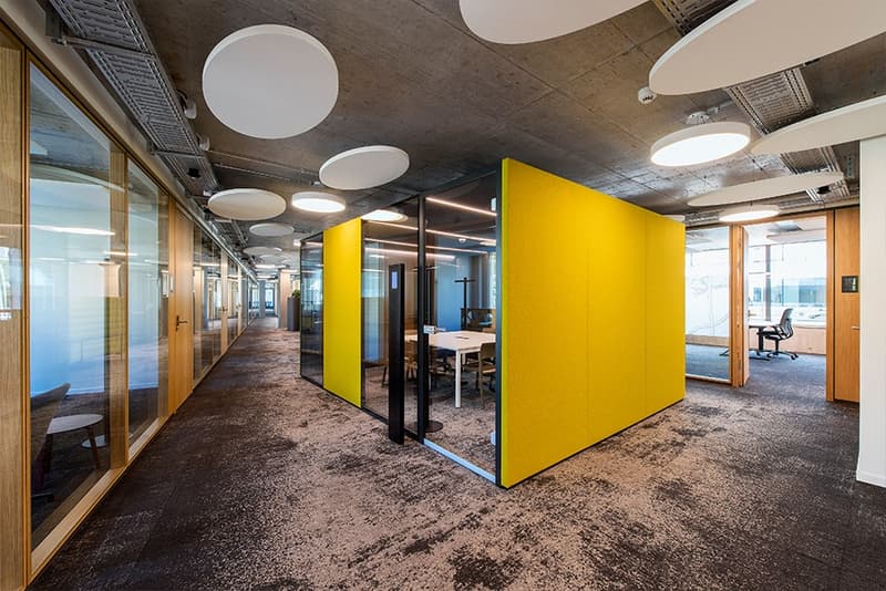 Open office layout with concrete ceiling, yellow accent walls, glass partitions, and modern lighting fixtures