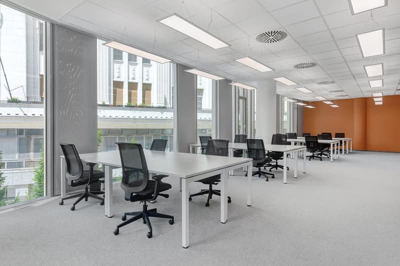 Large open office space with multiple workstations, white tables, black office chairs, and large windows providing natural light. The space has a modern and minimalist design with a mix of gray and orange accents.