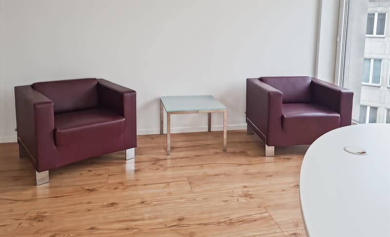 Two burgundy leather armchairs, a glass-topped coffee table, and a wooden floor in a bright, minimalist living room setting
