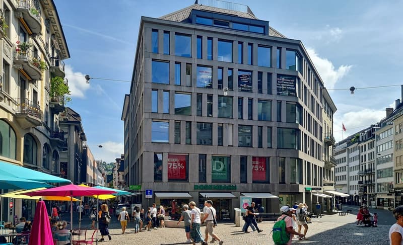 Modern building with glass windows, multiple floors, signage on the front, people walking around, street with shops and umbrellas