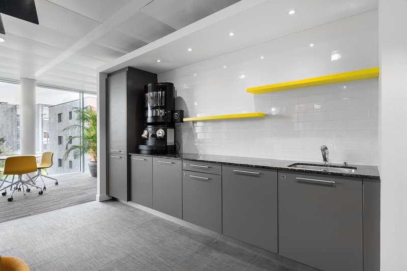 Modern kitchen with gray cabinets, black appliances, yellow accent shelves, and large windows providing natural light