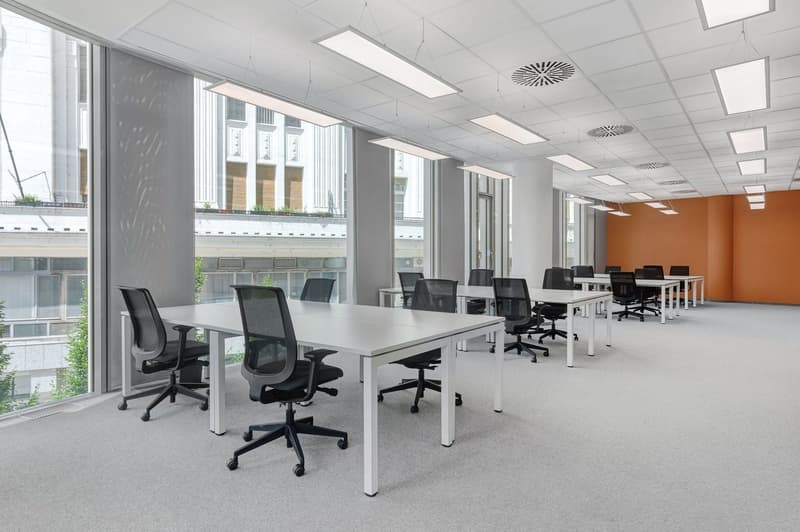 The image shows a modern, open-plan office space with large windows, white walls, and a gray carpet floor. The office is equipped with multiple workstations, each with a desk and an office chair. The desks are arranged in rows, and the space is well-lit b