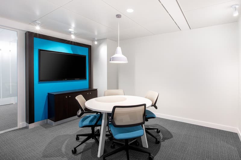 Modern office space with a large TV screen, a round white table, black chairs, and a cabinet. The room has white walls, gray carpeting, and recessed lighting.