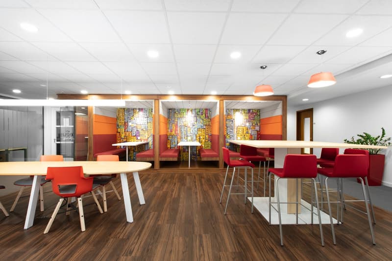 Open office space with colorful wall decor, wooden floors, round and rectangular tables, red and orange chairs, and a kitchen area with a dishwasher