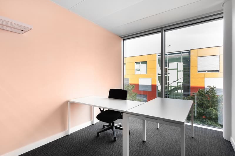 Spacious office with large windows, white desk, black office chair, and colorful exterior building visible through the windows
