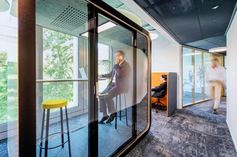 Modern office space with glass walls, high ceilings, and a view of the outdoor greenery. The space includes a bar-height table with a yellow stool, as well as private workspaces with chairs and desks.