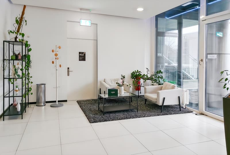 Spacious living room with large windows, white walls, tile flooring, modern furniture including a sofa and coffee table, and various potted plants and decor elements.