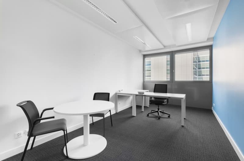 Spacious office room with white walls, large windows, black office chairs, a round white table, and a desk with a computer setup.