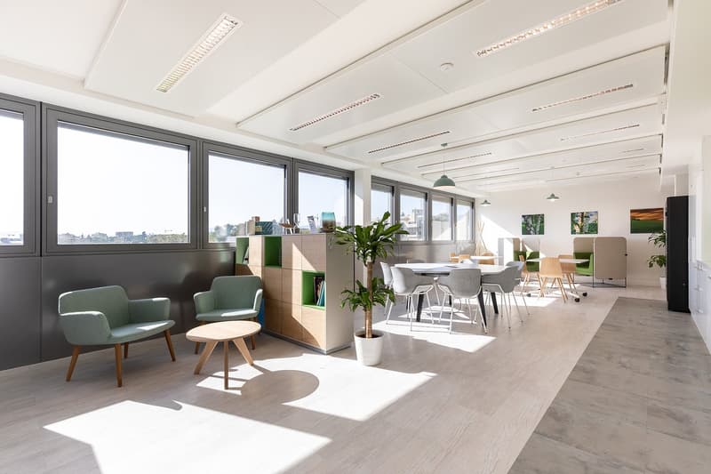 Spacious open-plan office with large windows providing a view, modern furniture including green armchairs and a dining table, storage units, and potted plants. The space has a bright and airy feel with a mix of work and relaxation areas.