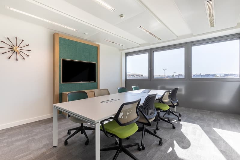Large conference room with a long table, comfortable office chairs, a TV mounted on the wall, and large windows providing a view of the surrounding area.