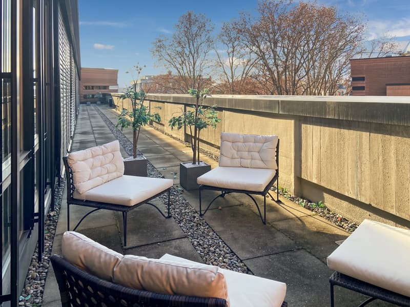 The image shows a cozy outdoor patio area with comfortable seating. There are two armchairs and a small side table on a paved surface, surrounded by potted plants and a concrete wall. The patio appears to be on a balcony or terrace, with a view of trees a