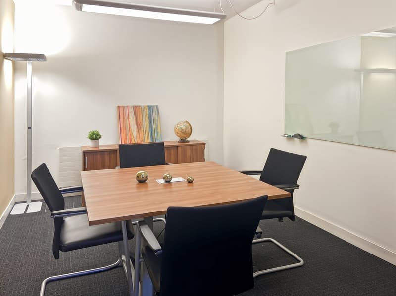 The image shows a modern and minimalist meeting room with a wooden table, black chairs, and various decor items such as a plant, artwork, and a globe. The room has white walls, recessed lighting, and a glass wall or window. There appears to be a built-in