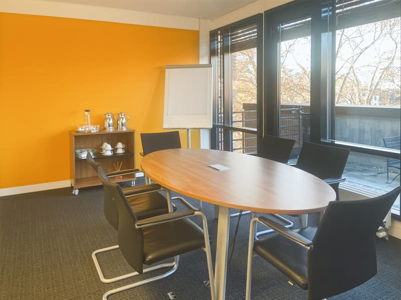 Bright, modern conference room with large windows, orange walls, wooden table, black chairs, and a whiteboard stand.