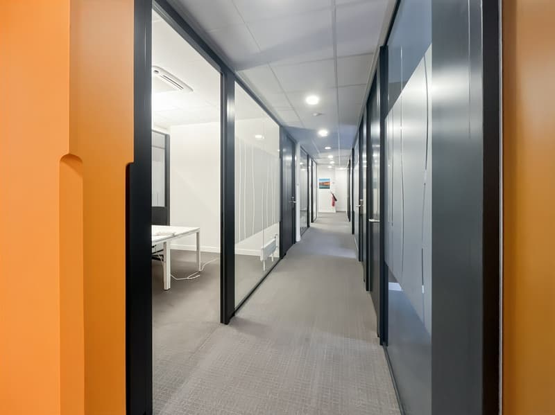 The image shows a long, well-lit office hallway with gray carpeted floors and white walls. The hallway has several doors on either side, likely leading to individual offices or workspaces. The lighting fixtures are recessed into the ceiling, providing eve