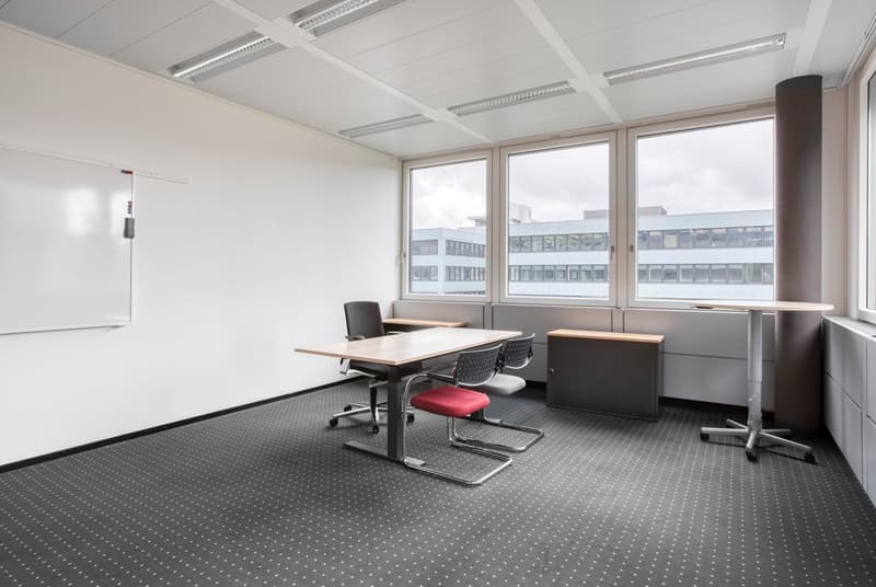 Spacious office room with large windows, white walls, suspended ceiling, desk, chairs, and storage cabinets