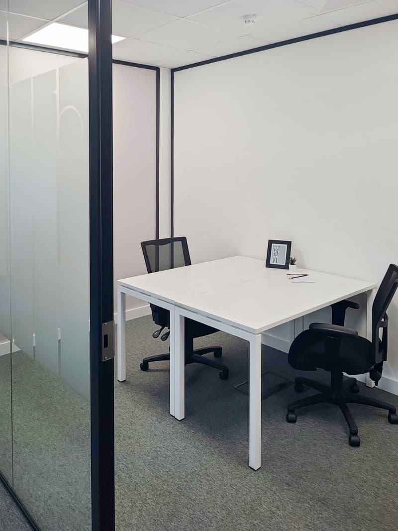 Modern office room with white desk and two black office chairs, framed picture on the desk, clear glass wall, grey carpet flooring