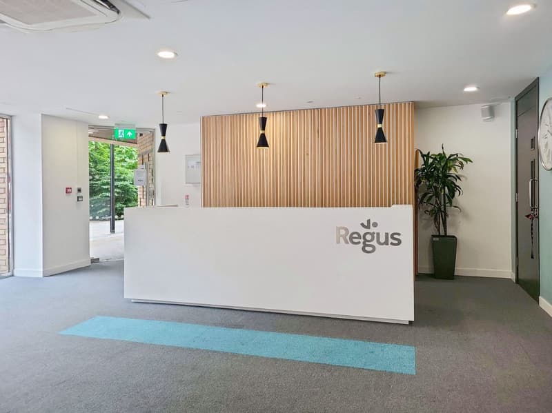 A modern lobby with a white reception desk, large windows, a potted plant, a wall with vertical slats, and three pendant lights. The floor has gray carpeting with a blue strip, and the wall is white with wooden slats and black lamps mounted on the ceiling