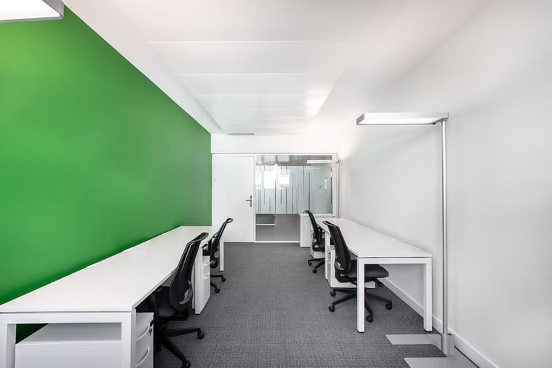 Open office space with white desks, black office chairs, and a green accent wall. The space has a modern and minimalist design with overhead lighting and carpeted flooring.