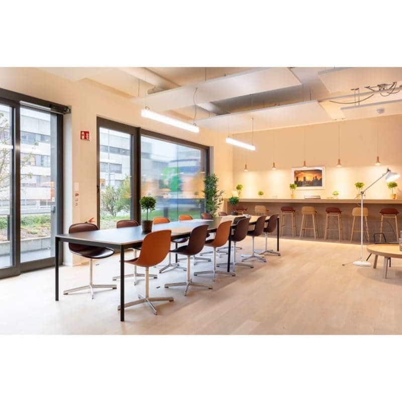 Large open office space with floor-to-ceiling windows, wooden floors, long conference table with chairs, bar-style seating area, and various potted plants throughout the room.