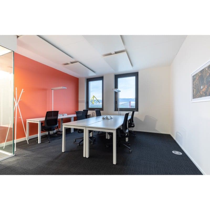 Modern office space with large windows, white walls, black chairs, and a long conference table. The room has a bright, open feel with a view of the outdoors.