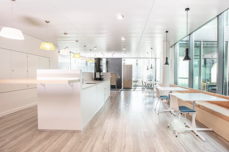 modern office space, white walls, wooden floor, white tables and chairs, kitchen area with sink and appliances, large windows, hanging lamps