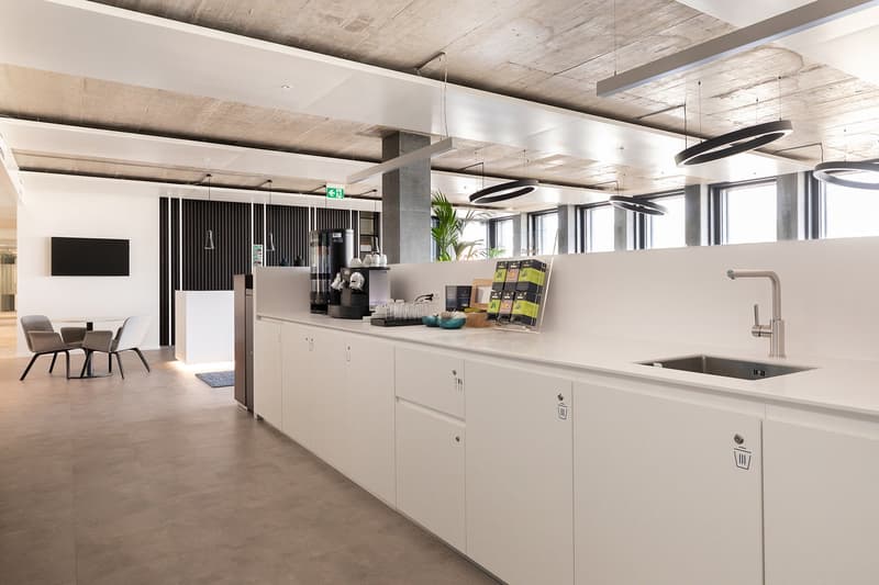 Minimalistic office design, white cabinets, under-mounted sink, modern lighting, a table with two chairs, a television on the wall, and coffee machines