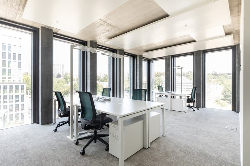 A modern office space with large windows, white desks, modern chairs, and gray carpeting.