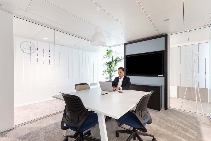 Office with meeting table, glass walls, a man, a laptop, a television on the wall, and a plant.