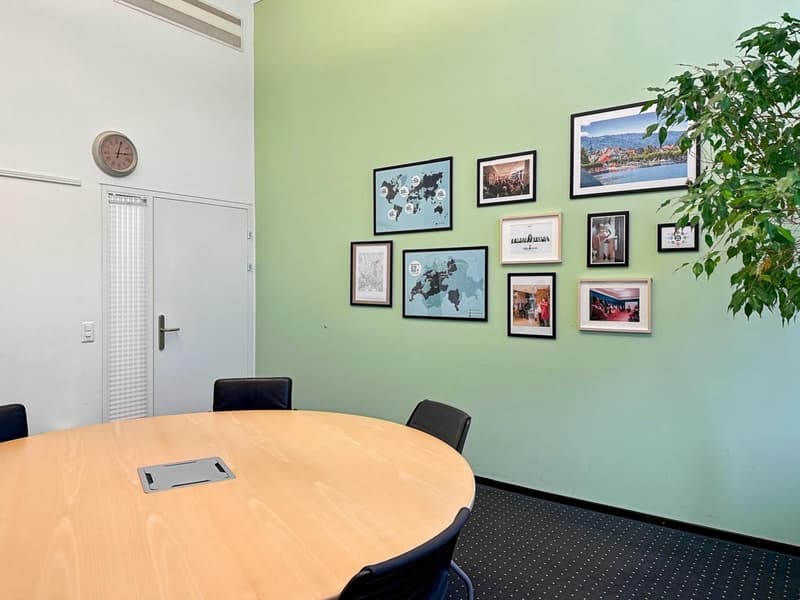 This image shows a small office space with a round wooden table, black chairs, and a wall decorated with various framed artwork and photographs. The room has a green accent wall and a plant in the corner, creating a cozy and professional atmosphere. There