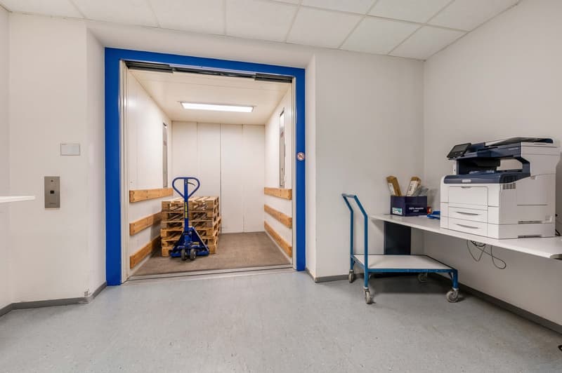 open door leading to storage area, forklift in doorway, copy machine on cart, pallet on floor, blue shelves on both sides of door