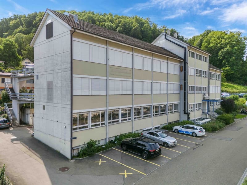 Multi-story office building, modern architecture, multiple cars parked, concrete parking lot, green surroundings