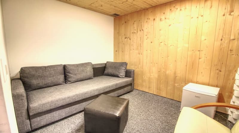 A small room with a gray sofa, a black ottoman, a white chair, and a wooden wall.
