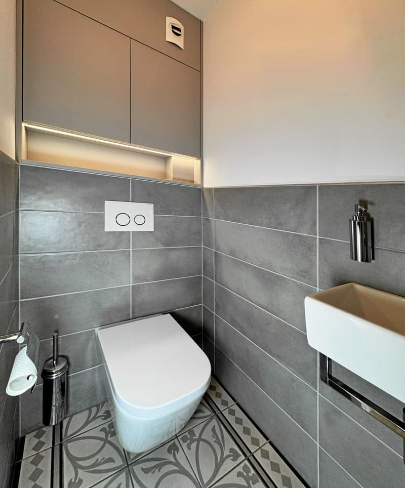 Modern small bathroom with tiled floor and walls, white sink and toilet, gray cabinet, soap dispenser