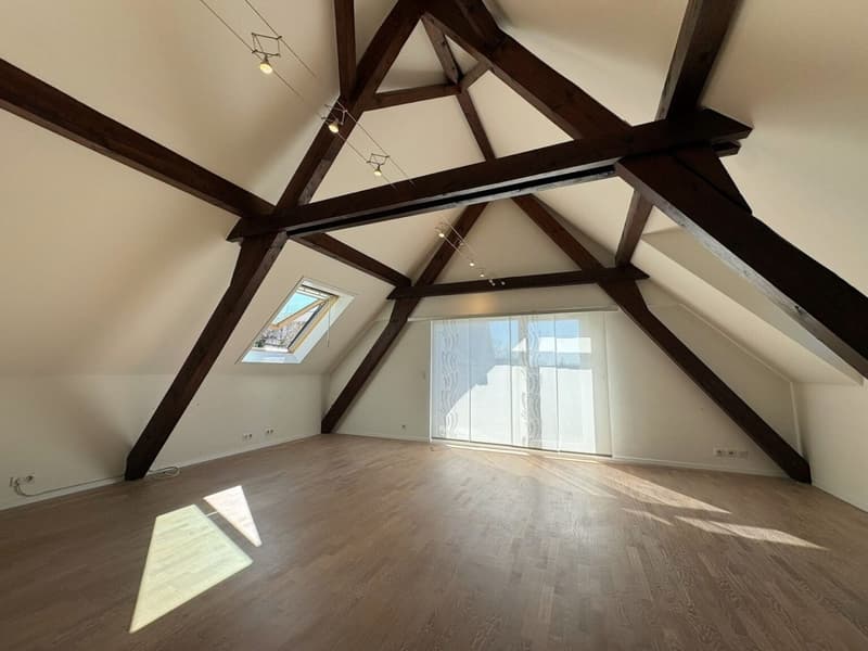 Empty attic room with wooden beams, large windows, white walls, wooden floor, ceiling lights