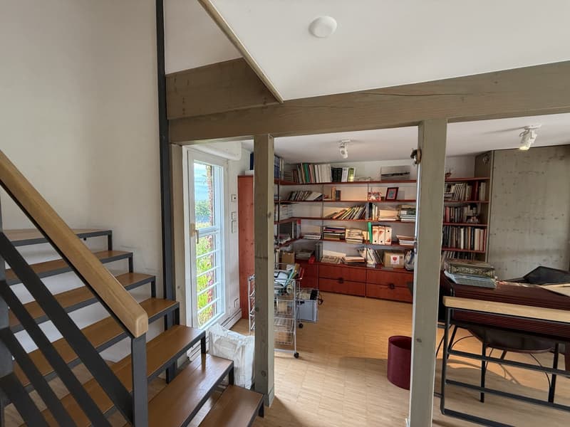 Interior with wooden stairs, wooden floor, shelves filled with books and items, table with chair, exit door, and light fixtures