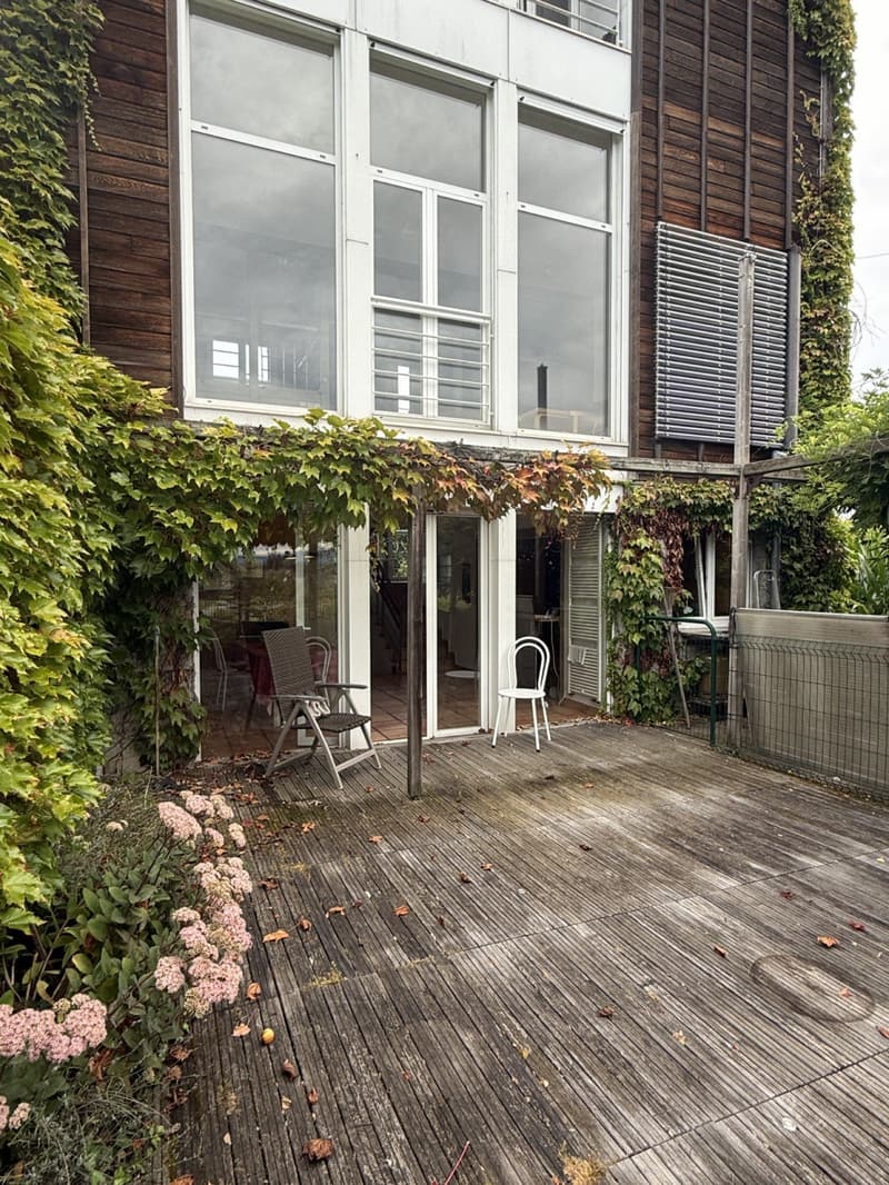 Raised wooden deck, white chairs, wooden and white walls, large windows, vine-covered trellis, small garden area