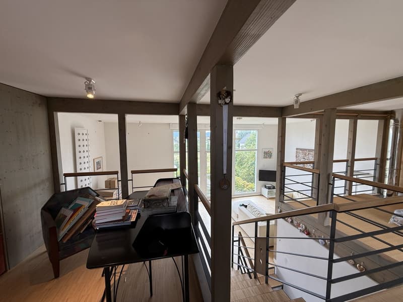 Living room with a table, chair, and stairs, elevated ground floor, exposed wooden beams, open layout, large windows, metal railings, and an upper floor