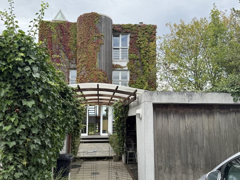 Curved house covered in greenery, main entrance with a wooden walkway, attached garage, balcony visible above.