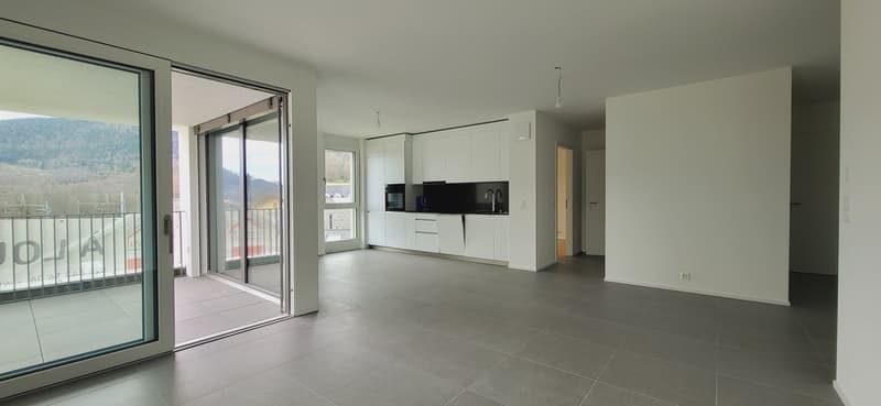 Open-concept living space with large windows, sliding glass doors leading to a balcony, modern white kitchen with black countertops, and tiled flooring throughout.
