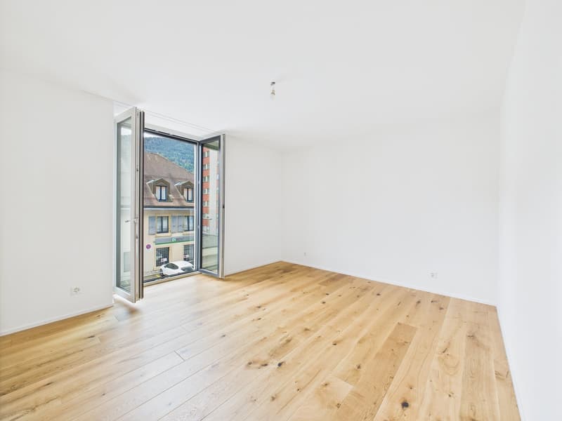 Spacious room with large windows, hardwood flooring, and a balcony or terrace overlooking the surrounding buildings.