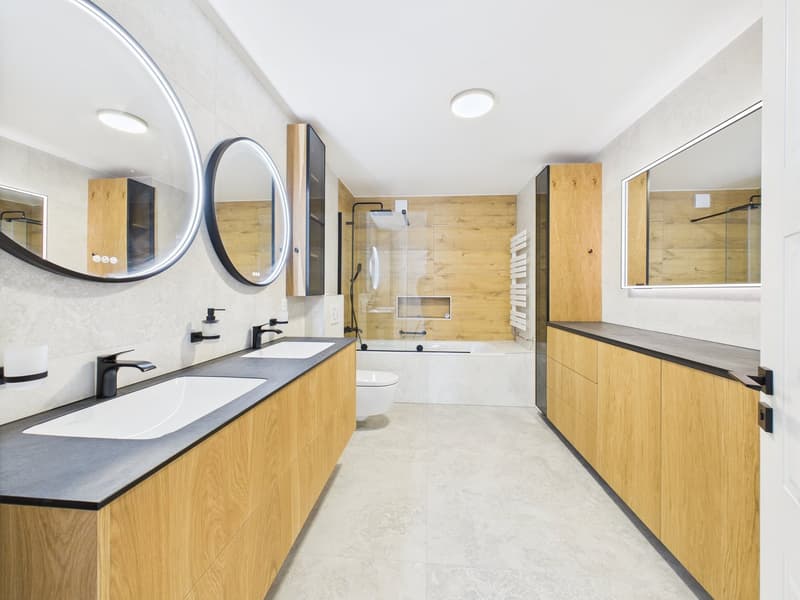 Spacious bathroom with wood-paneled walls, double vanity sinks, large round mirrors, and a glass-enclosed shower area