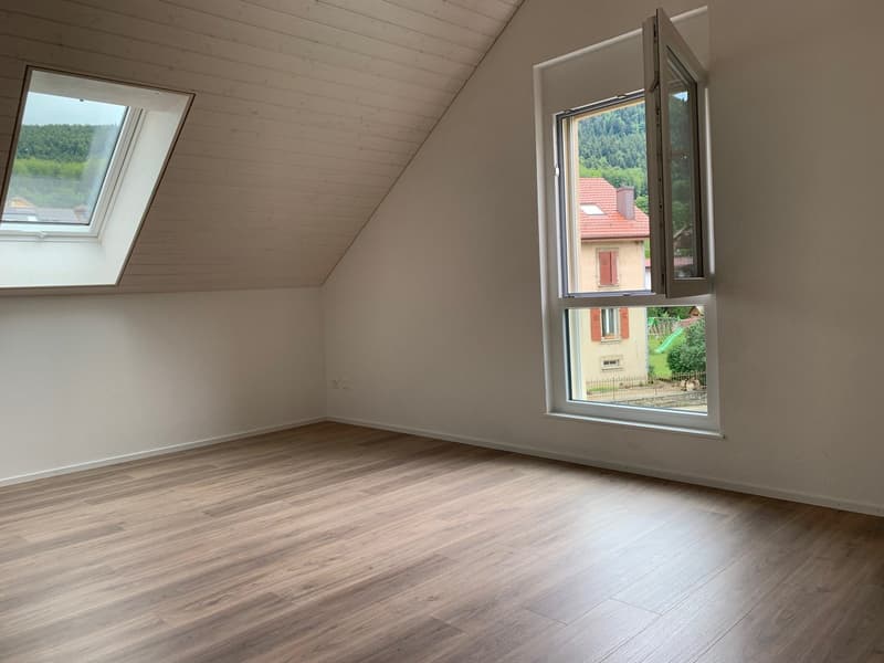 An empty room with wooden flooring, two windows, and a sloping ceiling, overlooking a house and a mountain range
